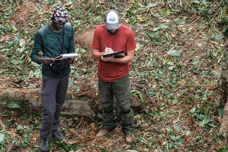Two men stand side by side in a forested area. Both are writing on notepads held in their hands.